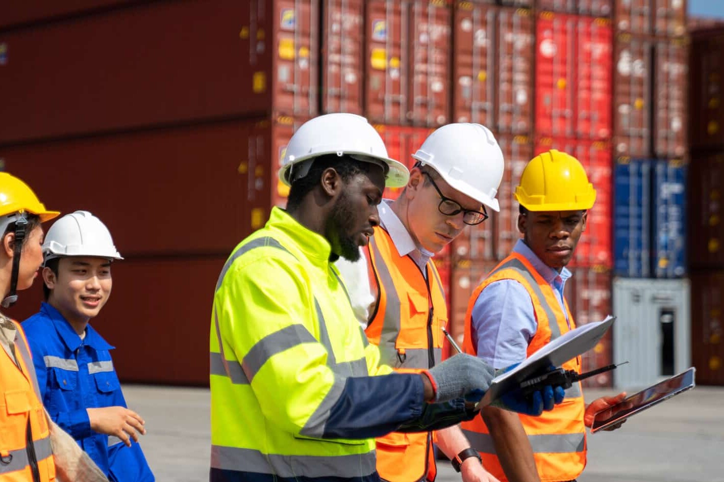A diverse group of workers in safety gear and hard hats review documents outdoors at a shipping container yard, as stacked red containers loom in the background amid trade wars as pessimism about economy deepens.