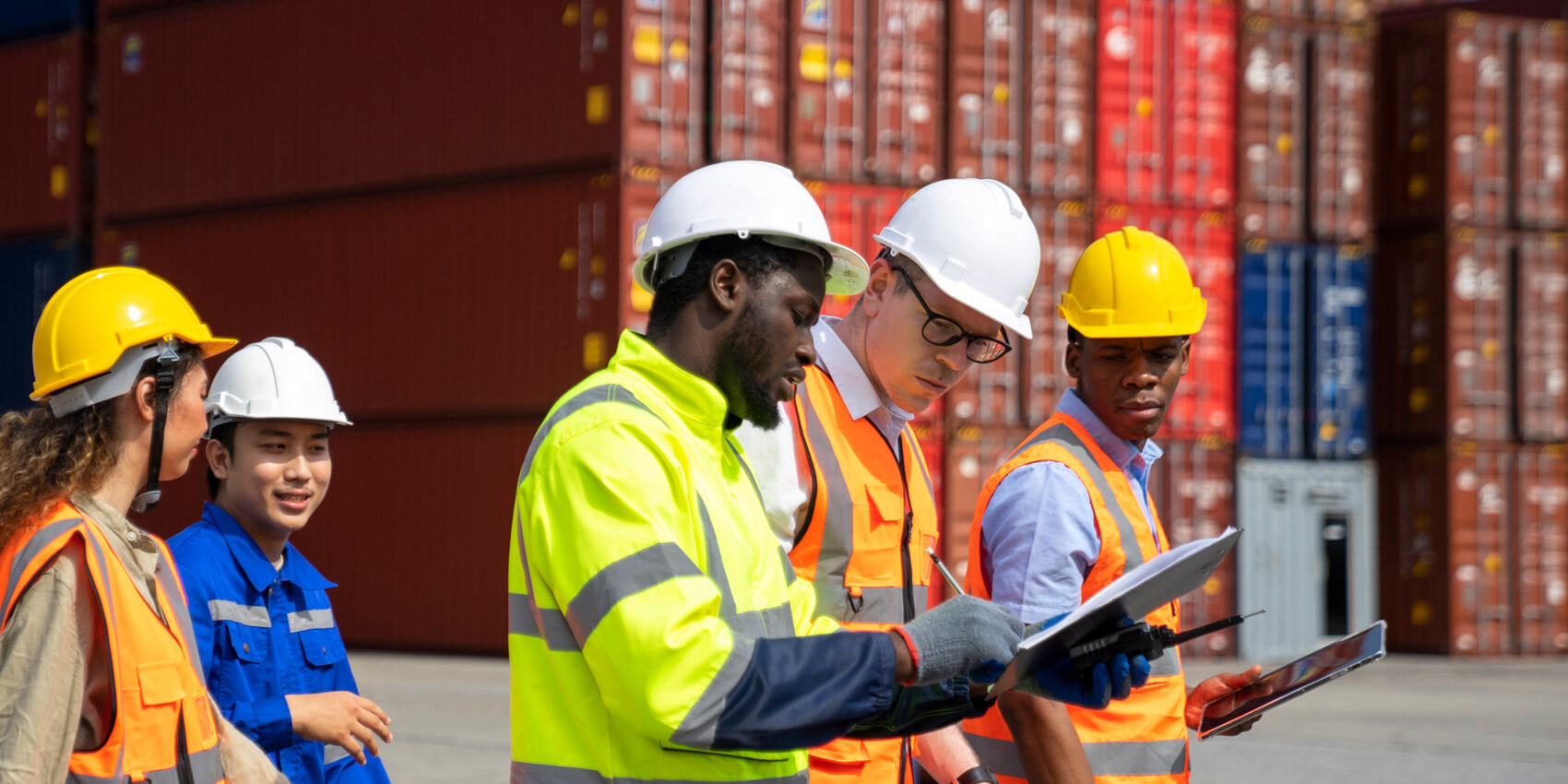 A diverse group of workers in safety gear and hard hats review documents outdoors at a shipping container yard, as stacked red containers loom in the background amid trade wars as pessimism about economy deepens.