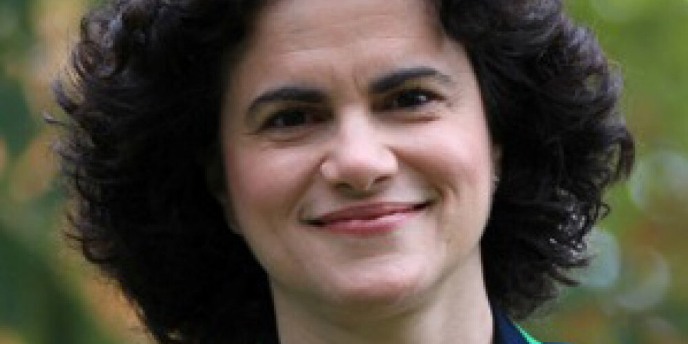 Michael Mendelson, with short, curly dark hair, smiles outdoors. Wearing a navy blazer with green trim and a delicate necklace, they stand against a backdrop of blurred greenery.