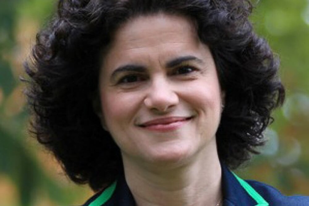Michael Mendelson, with short, curly dark hair, smiles outdoors. Wearing a navy blazer with green trim and a delicate necklace, they stand against a backdrop of blurred greenery.