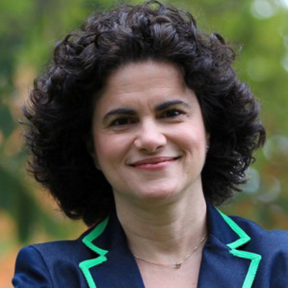 Michael Mendelson, with short, curly dark hair, smiles outdoors. Wearing a navy blazer with green trim and a delicate necklace, they stand against a backdrop of blurred greenery.