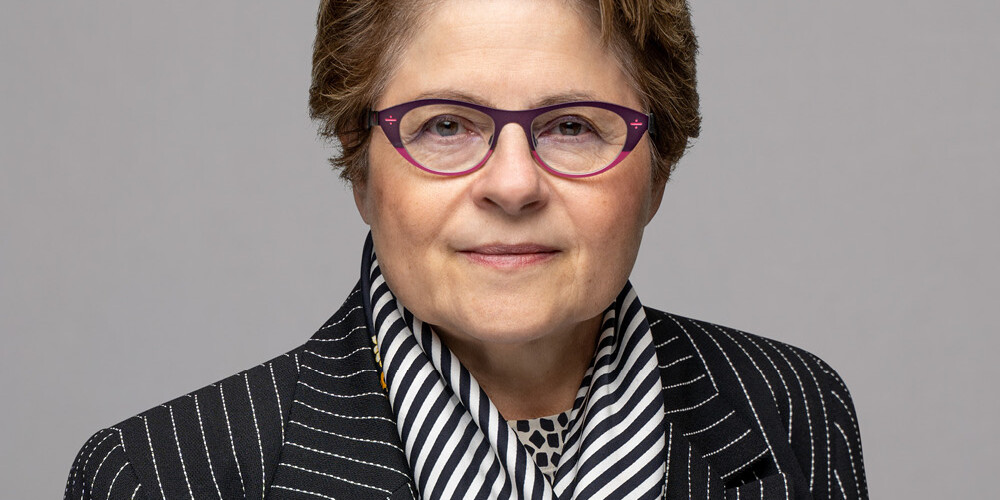 Michael Mendelson, with short light brown hair, purple glasses, a black pinstripe blazer, black and white striped scarf, and a patterned shirt, poses confidently in front of a plain gray background.