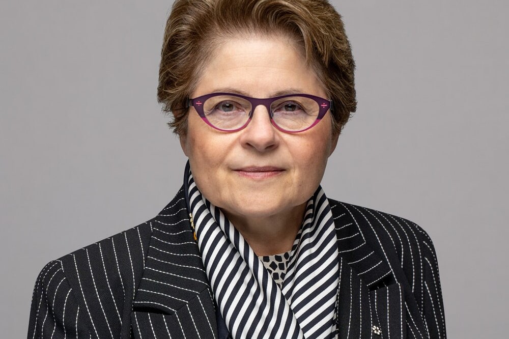 Michael Mendelson, with short light brown hair, purple glasses, a black pinstripe blazer, black and white striped scarf, and a patterned shirt, poses confidently in front of a plain gray background.