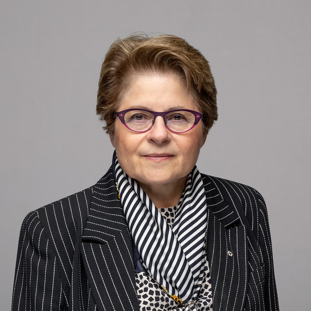 Michael Mendelson, with short light brown hair, purple glasses, a black pinstripe blazer, black and white striped scarf, and a patterned shirt, poses confidently in front of a plain gray background.