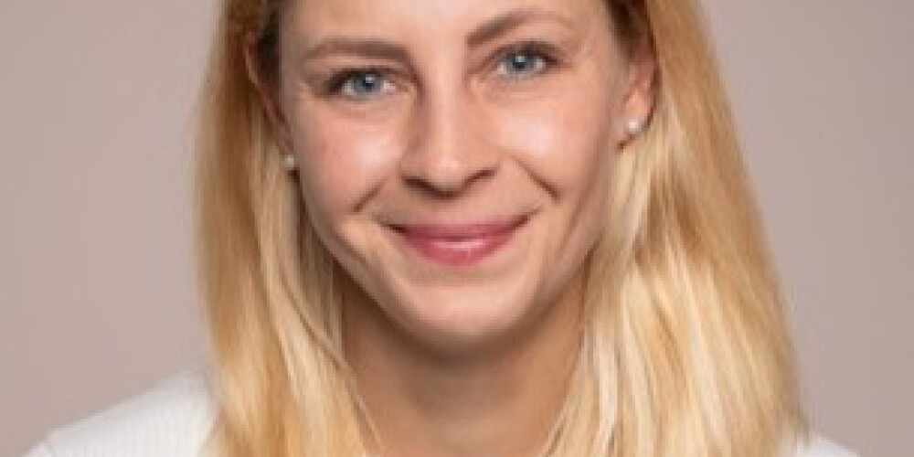 A woman with straight, shoulder-length blonde hair and blue eyes smiles at the camera. She is wearing a white sweater and is posed in front of a plain, light-colored background, reminiscent of a portrait by Michael Mendelson.