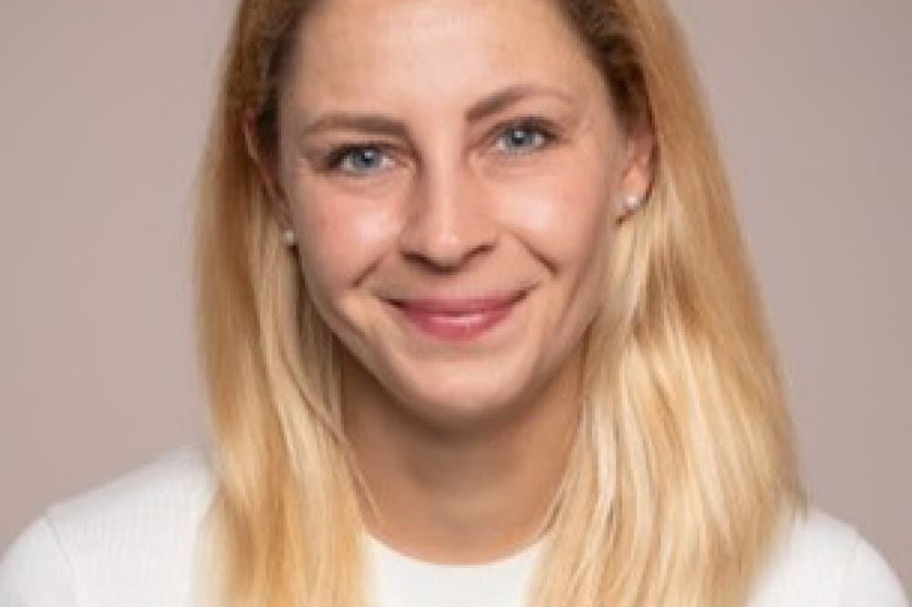 A woman with straight, shoulder-length blonde hair and blue eyes smiles at the camera. She is wearing a white sweater and is posed in front of a plain, light-colored background, reminiscent of a portrait by Michael Mendelson.