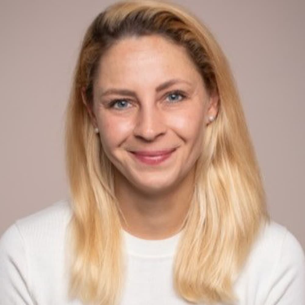 A woman with straight, shoulder-length blonde hair and blue eyes smiles at the camera. She is wearing a white sweater and is posed in front of a plain, light-colored background, reminiscent of a portrait by Michael Mendelson.