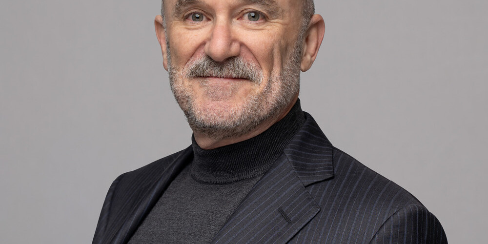 Michael Mendelson, a bald man with a gray beard, wears a dark pinstripe suit and charcoal turtleneck, smiling slightly as he stands against a plain light gray background.