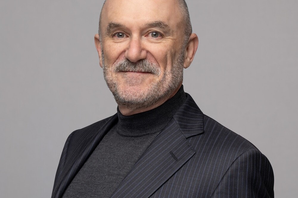 Michael Mendelson, a bald man with a gray beard, wears a dark pinstripe suit and charcoal turtleneck, smiling slightly as he stands against a plain light gray background.
