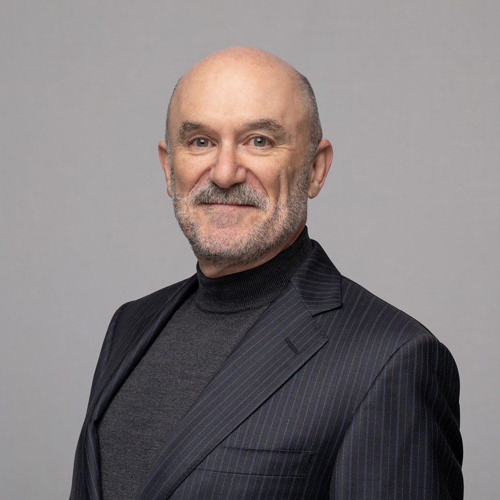 Michael Mendelson, a bald man with a gray beard, wears a dark pinstripe suit and charcoal turtleneck, smiling slightly as he stands against a plain light gray background.