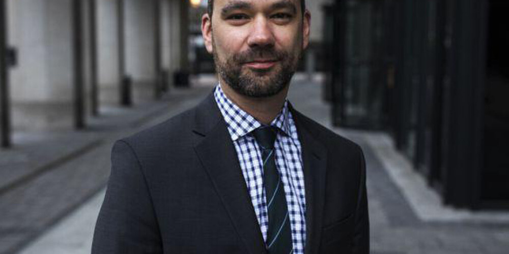 Michael Mendelson stands on a city sidewalk in a dark suit, checkered shirt, and striped tie. Tall buildings and glass windows line the street behind him as he faces the camera and smiles slightly.