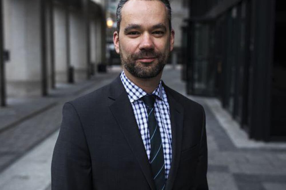 Michael Mendelson stands on a city sidewalk in a dark suit, checkered shirt, and striped tie. Tall buildings and glass windows line the street behind him as he faces the camera and smiles slightly.