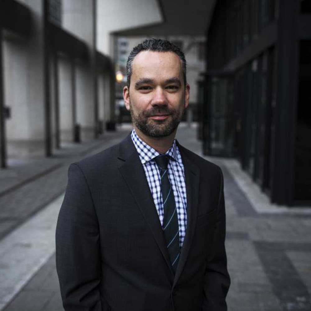 Michael Mendelson stands on a city sidewalk in a dark suit, checkered shirt, and striped tie. Tall buildings and glass windows line the street behind him as he faces the camera and smiles slightly.