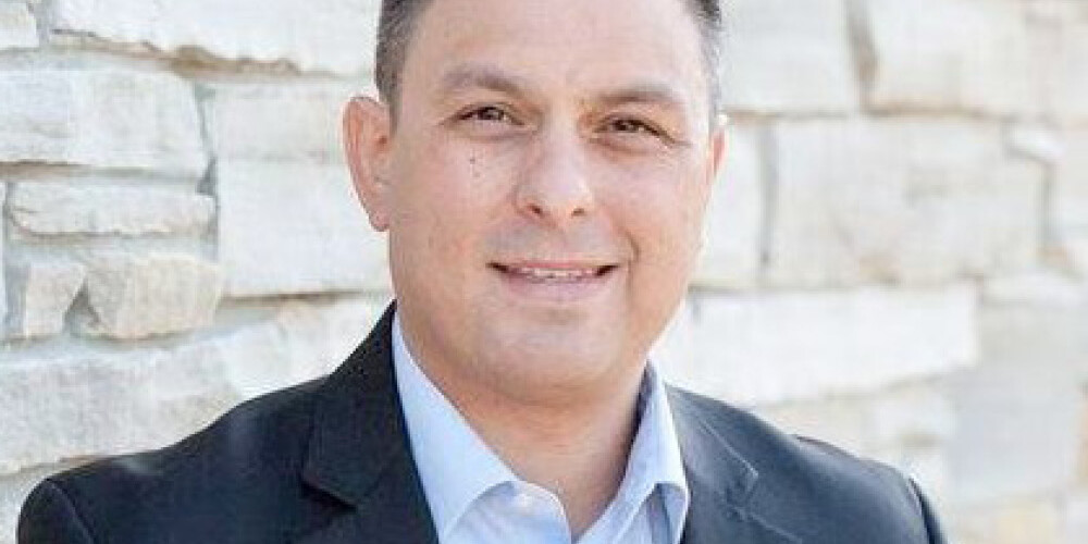 Michael Mendelson, a man with short dark hair wearing a black suit jacket and light blue shirt, stands smiling in front of a light stone wall.