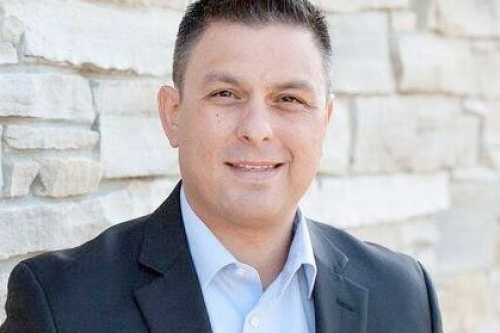 Michael Mendelson, a man with short dark hair wearing a black suit jacket and light blue shirt, stands smiling in front of a light stone wall.
