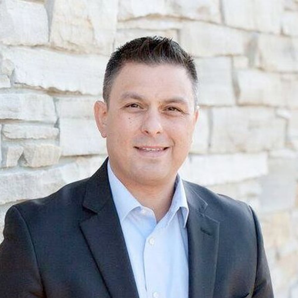 Michael Mendelson, a man with short dark hair wearing a black suit jacket and light blue shirt, stands smiling in front of a light stone wall.