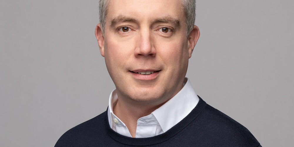 Michael Mendelson, a man with short gray hair, wears a white collared shirt under a dark sweater, posing and smiling softly against a plain light gray background.