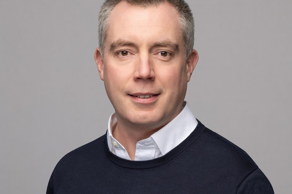 Michael Mendelson, a man with short gray hair, wears a white collared shirt under a dark sweater, posing and smiling softly against a plain light gray background.