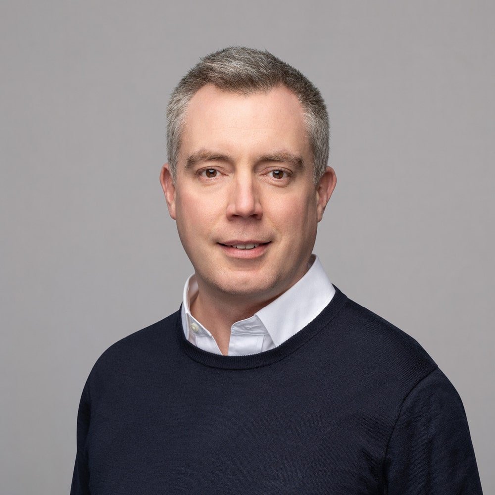 Michael Mendelson, a man with short gray hair, wears a white collared shirt under a dark sweater, posing and smiling softly against a plain light gray background.