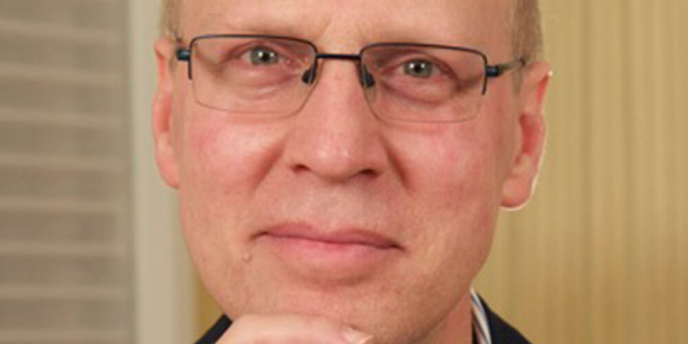 Andrew Griffith, a middle-aged person with glasses, short light hair, and a thoughtful expression rests their chin on their hand, wearing a dark jacket and striped shirt. The background features vertical and horizontal patterns.