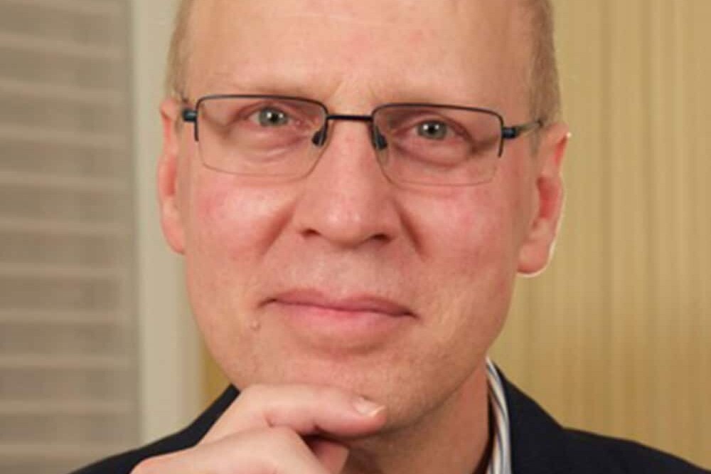 Andrew Griffith, a middle-aged person with glasses, short light hair, and a thoughtful expression rests their chin on their hand, wearing a dark jacket and striped shirt. The background features vertical and horizontal patterns.