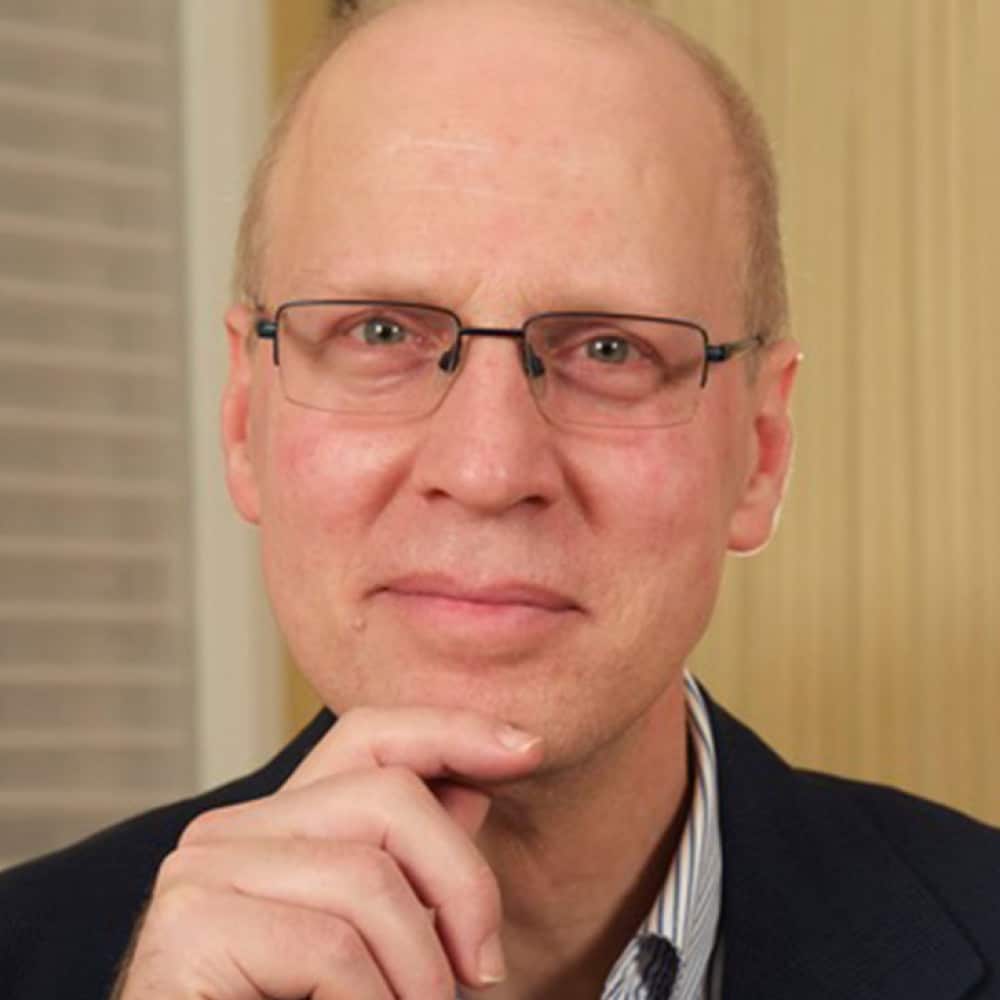 Andrew Griffith, a middle-aged person with glasses, short light hair, and a thoughtful expression rests their chin on their hand, wearing a dark jacket and striped shirt. The background features vertical and horizontal patterns.