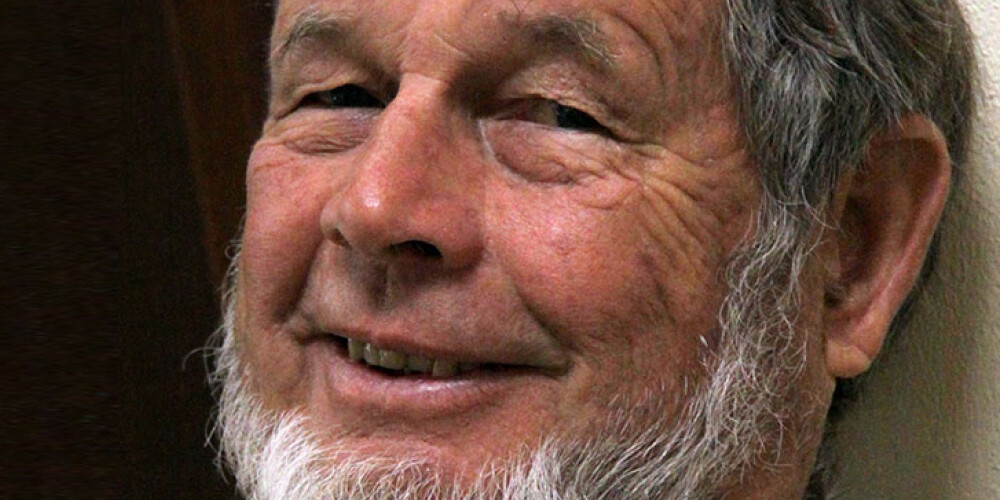 Andrew Griffith, an older man with a short white beard and gray hair, smiles gently while leaning against a wall, wearing a tan jacket and a beaded necklace.