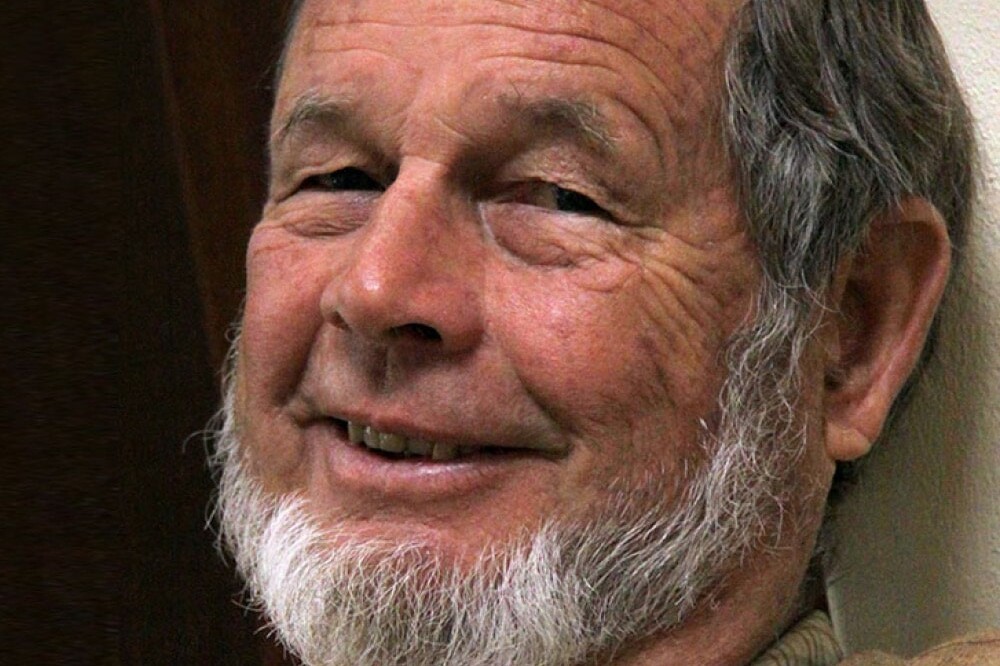 Andrew Griffith, an older man with a short white beard and gray hair, smiles gently while leaning against a wall, wearing a tan jacket and a beaded necklace.