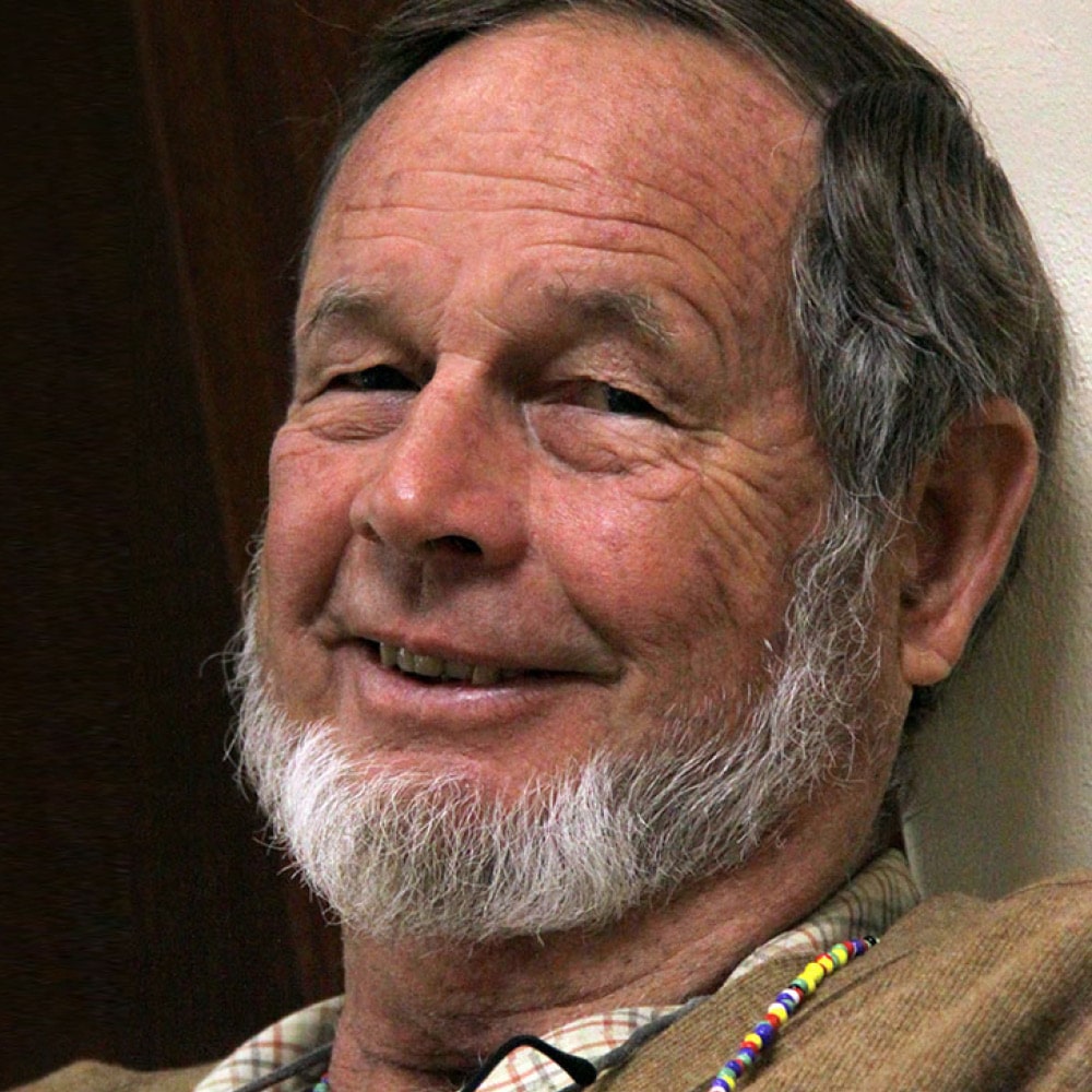Andrew Griffith, an older man with a short white beard and gray hair, smiles gently while leaning against a wall, wearing a tan jacket and a beaded necklace.