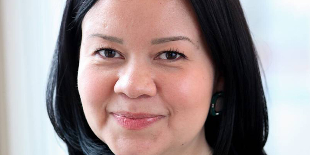 A woman with straight black hair, wearing a black blazer and patterned top, smiles softly at the camera. She is standing indoors in front of a bright window in Andrew Griffith’s office.