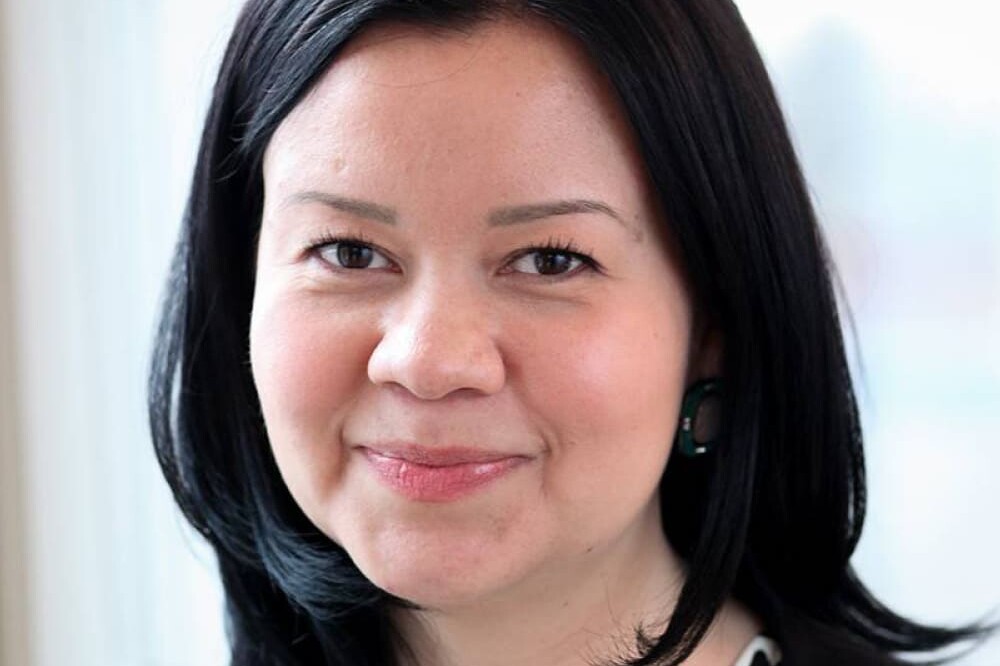 A woman with straight black hair, wearing a black blazer and patterned top, smiles softly at the camera. She is standing indoors in front of a bright window in Andrew Griffith’s office.