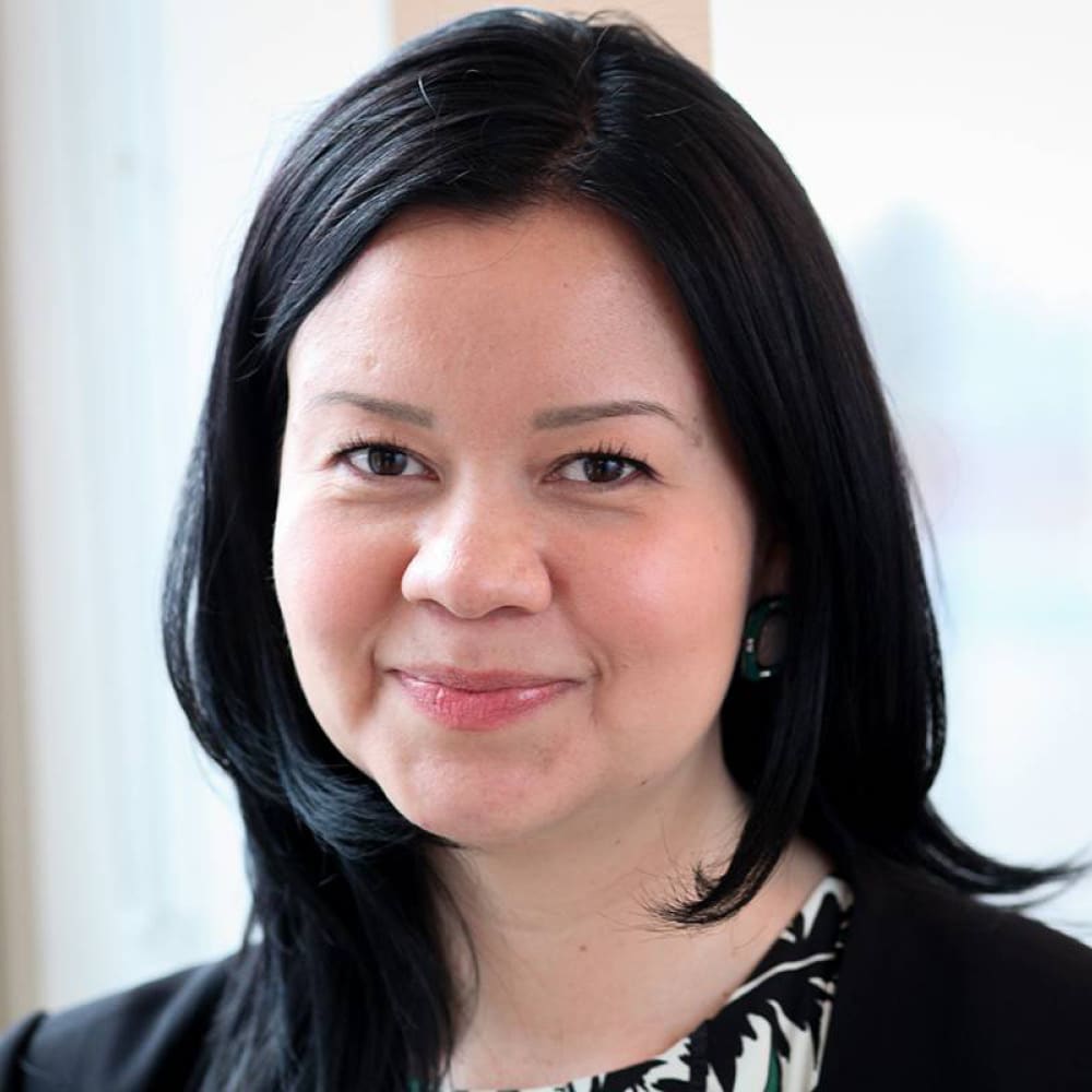 A woman with straight black hair, wearing a black blazer and patterned top, smiles softly at the camera. She is standing indoors in front of a bright window in Andrew Griffith’s office.