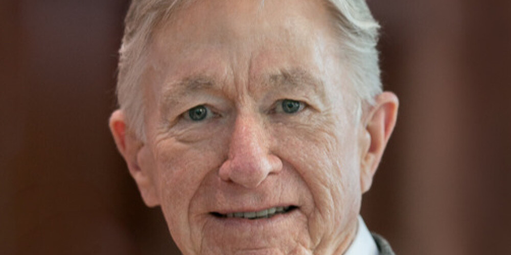 Andrew Griffith, an older man with gray hair, smiles gently at the camera. He is wearing a white collared shirt and a gray blazer. The background is softly blurred in shades of brown.