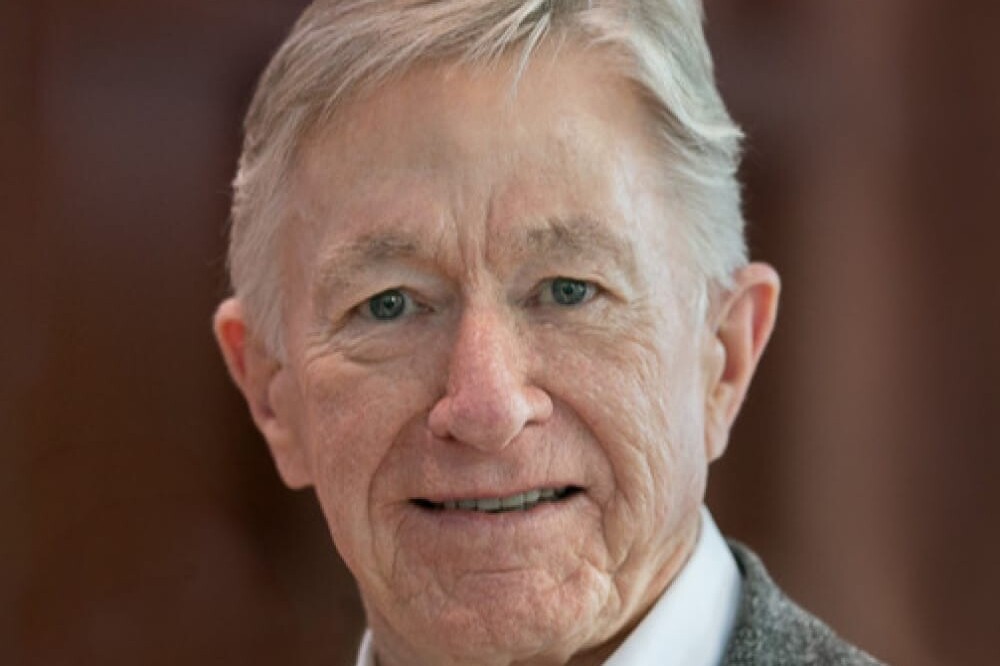 Andrew Griffith, an older man with gray hair, smiles gently at the camera. He is wearing a white collared shirt and a gray blazer. The background is softly blurred in shades of brown.