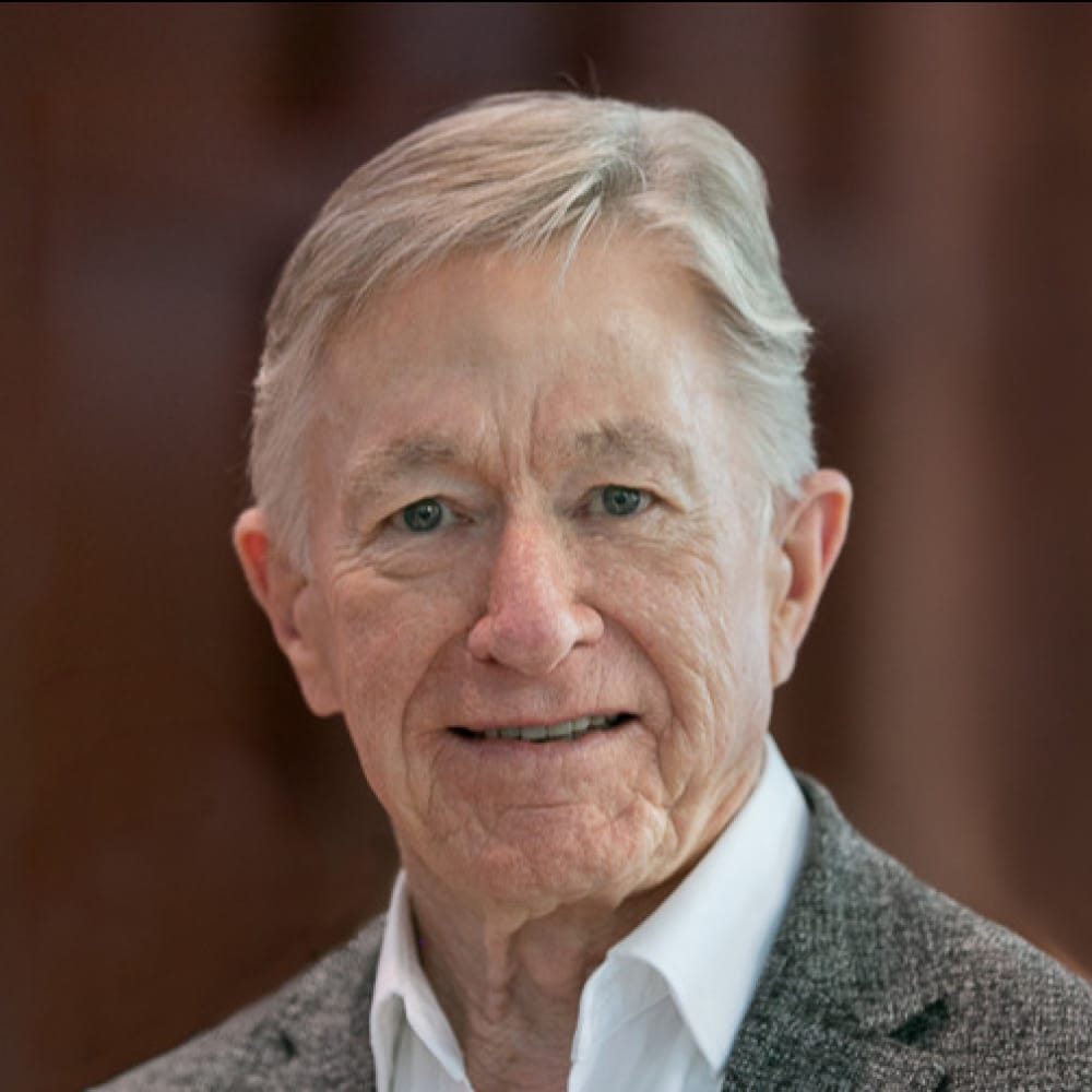Andrew Griffith, an older man with gray hair, smiles gently at the camera. He is wearing a white collared shirt and a gray blazer. The background is softly blurred in shades of brown.