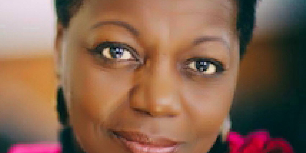 A woman with short hair smiles softly, wearing a vibrant red textured scarf. The background is blurred, drawing attention to her expressive eyes and warm expression—captured in the distinctive style of Andrew Griffith.