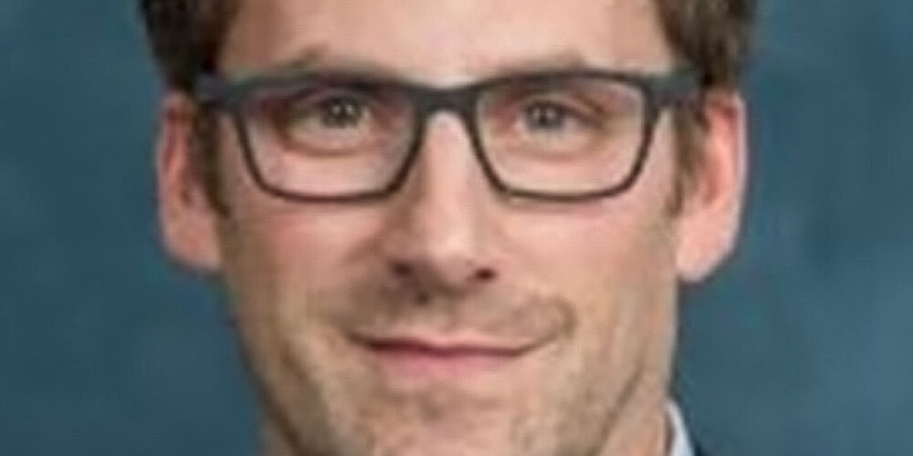 Andrew Griffith, a man with short brown hair and glasses, wearing a blue suit jacket and light blue collared shirt, poses in front of a plain blue background.