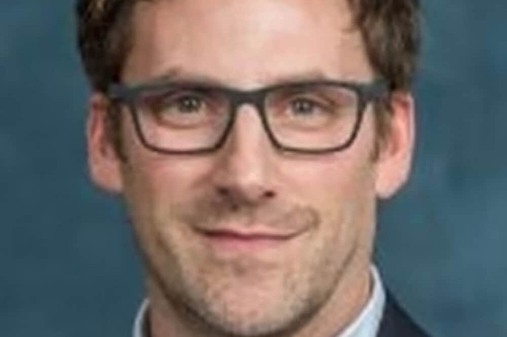 Andrew Griffith, a man with short brown hair and glasses, wearing a blue suit jacket and light blue collared shirt, poses in front of a plain blue background.
