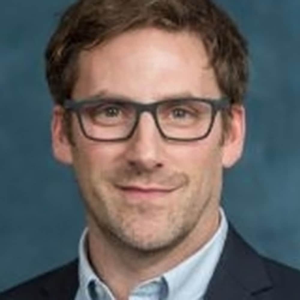 Andrew Griffith, a man with short brown hair and glasses, wearing a blue suit jacket and light blue collared shirt, poses in front of a plain blue background.