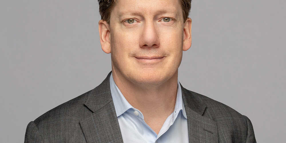 Andrew Parkin, a man with light brown hair, wears a gray suit jacket and light blue shirt as he poses against a plain light gray background, looking at the camera with a neutral expression.