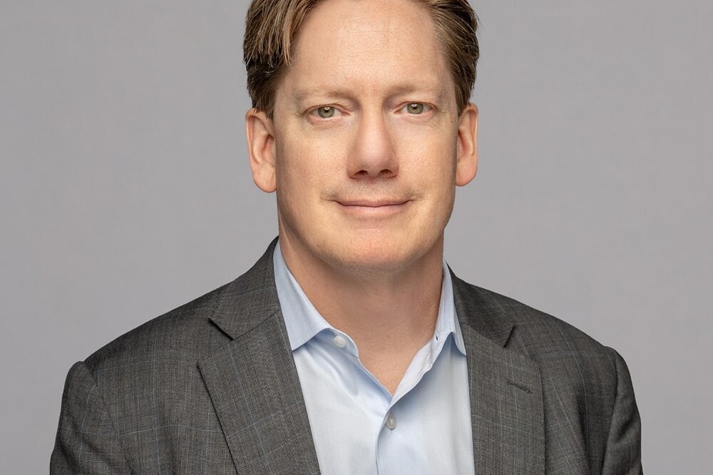 Andrew Parkin, a man with light brown hair, wears a gray suit jacket and light blue shirt as he poses against a plain light gray background, looking at the camera with a neutral expression.