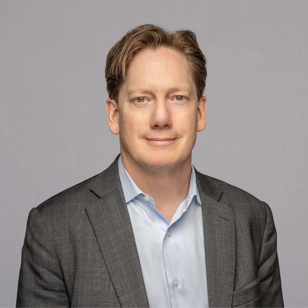 Andrew Parkin, a man with light brown hair, wears a gray suit jacket and light blue shirt as he poses against a plain light gray background, looking at the camera with a neutral expression.