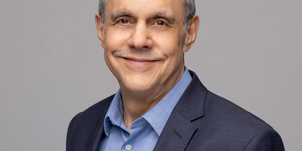 Andrew Parkin, a middle-aged man with short gray hair, smiles while wearing a blue dress shirt and dark blazer against a plain light gray background.