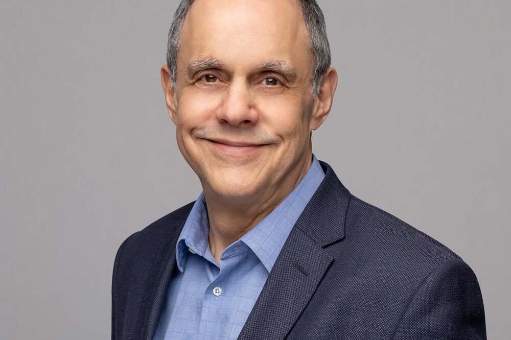 Andrew Parkin, a middle-aged man with short gray hair, smiles while wearing a blue dress shirt and dark blazer against a plain light gray background.