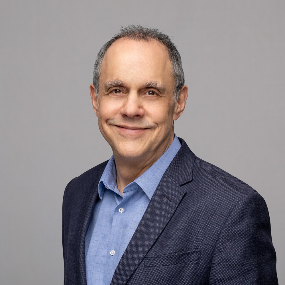 Andrew Parkin, a middle-aged man with short gray hair, smiles while wearing a blue dress shirt and dark blazer against a plain light gray background.