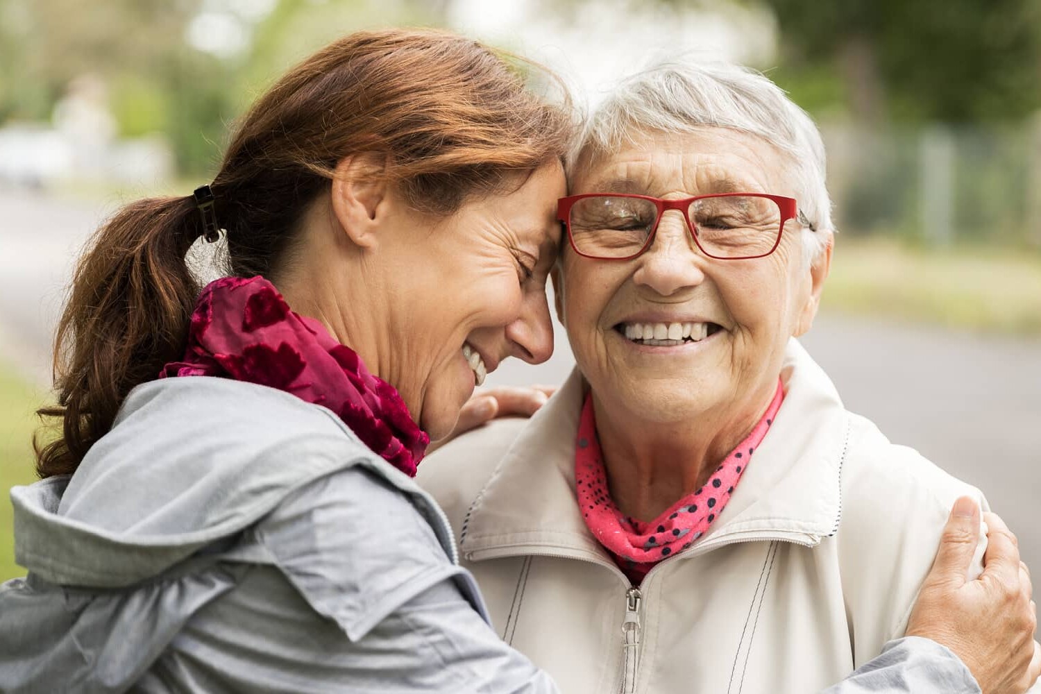 Two women, one older and one younger, share a joyful embrace outdoors. The older woman wears glasses and a light jacket; both wear pink scarves and smile warmly. Our elbows may be up but have Canadians really changed? Trees and a path are visible in the background.