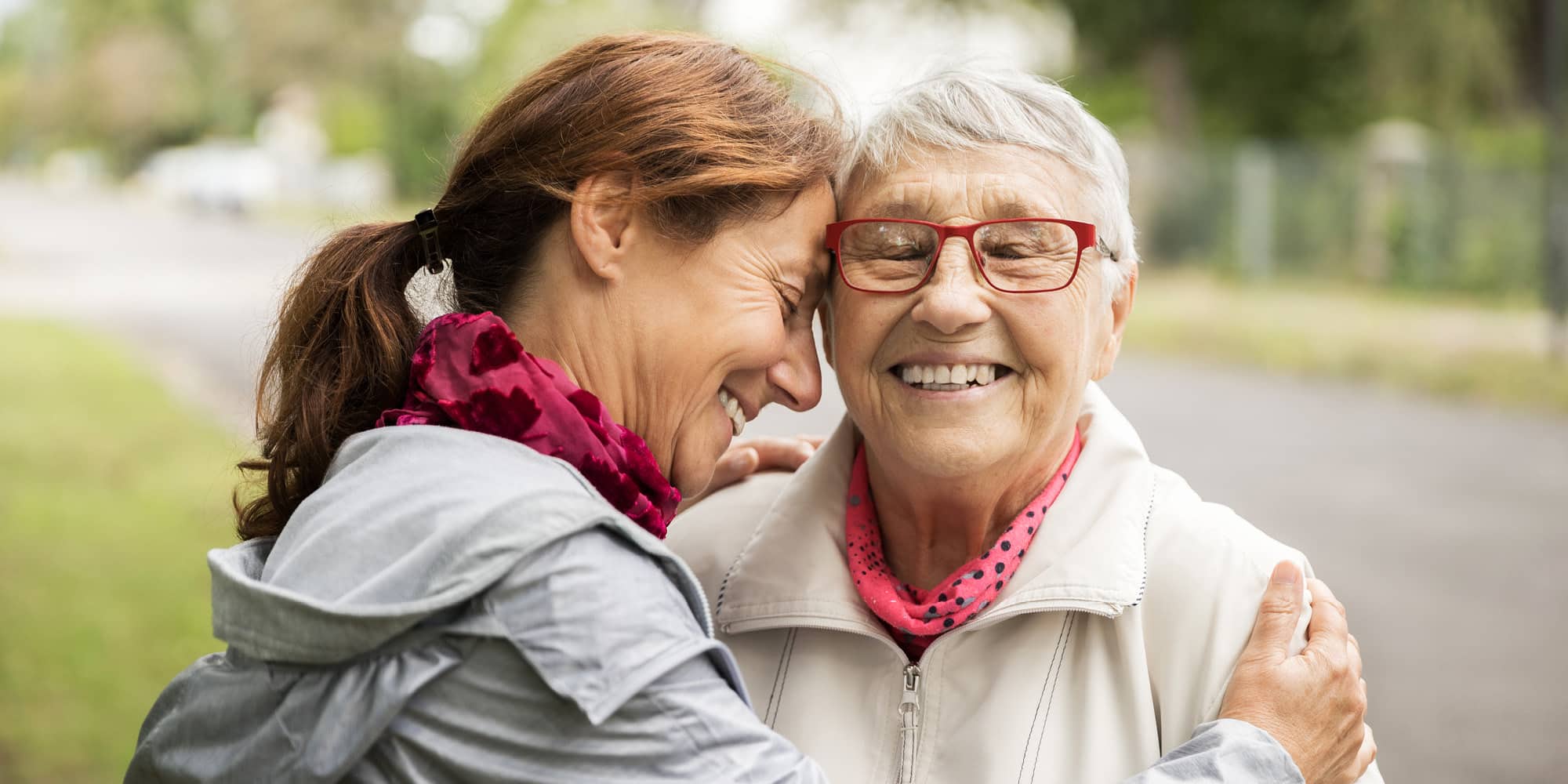 Two women, one older and one younger, share a joyful embrace outdoors. The older woman wears glasses and a light jacket; both wear pink scarves and smile warmly. Our elbows may be up but have Canadians really changed? Trees and a path are visible in the background.