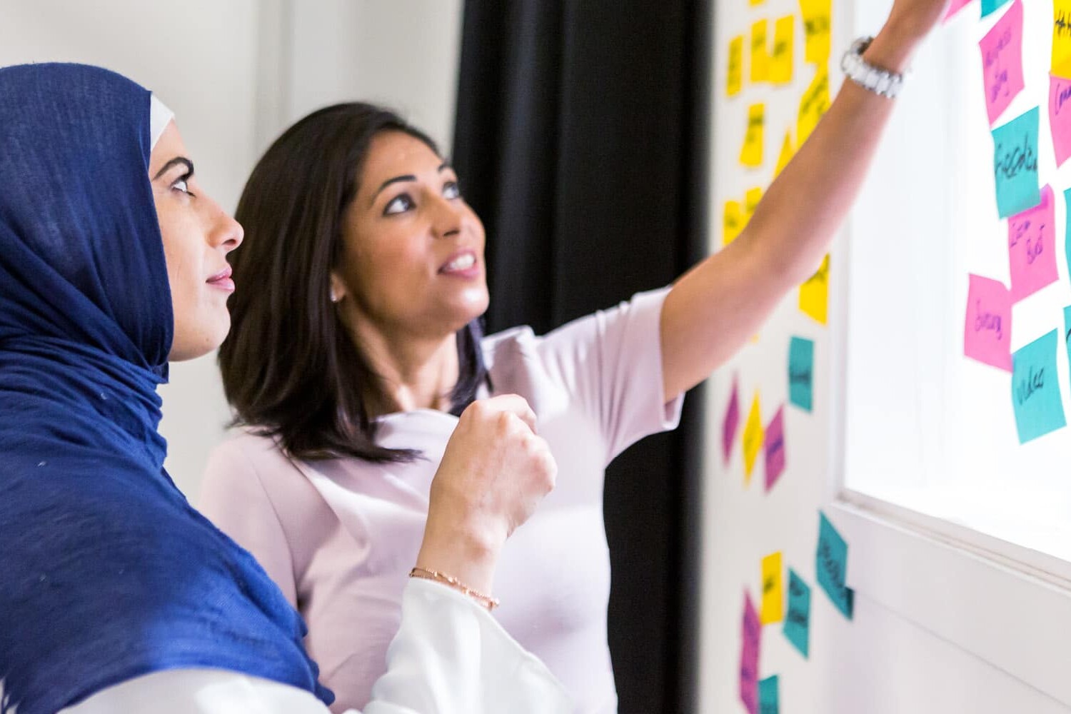 Two women stand by a window covered with colorful sticky notes, discussing and pointing at the notes. In this skills training Canada session, one woman wears a blue hijab, and both appear focused and engaged in collaboration.