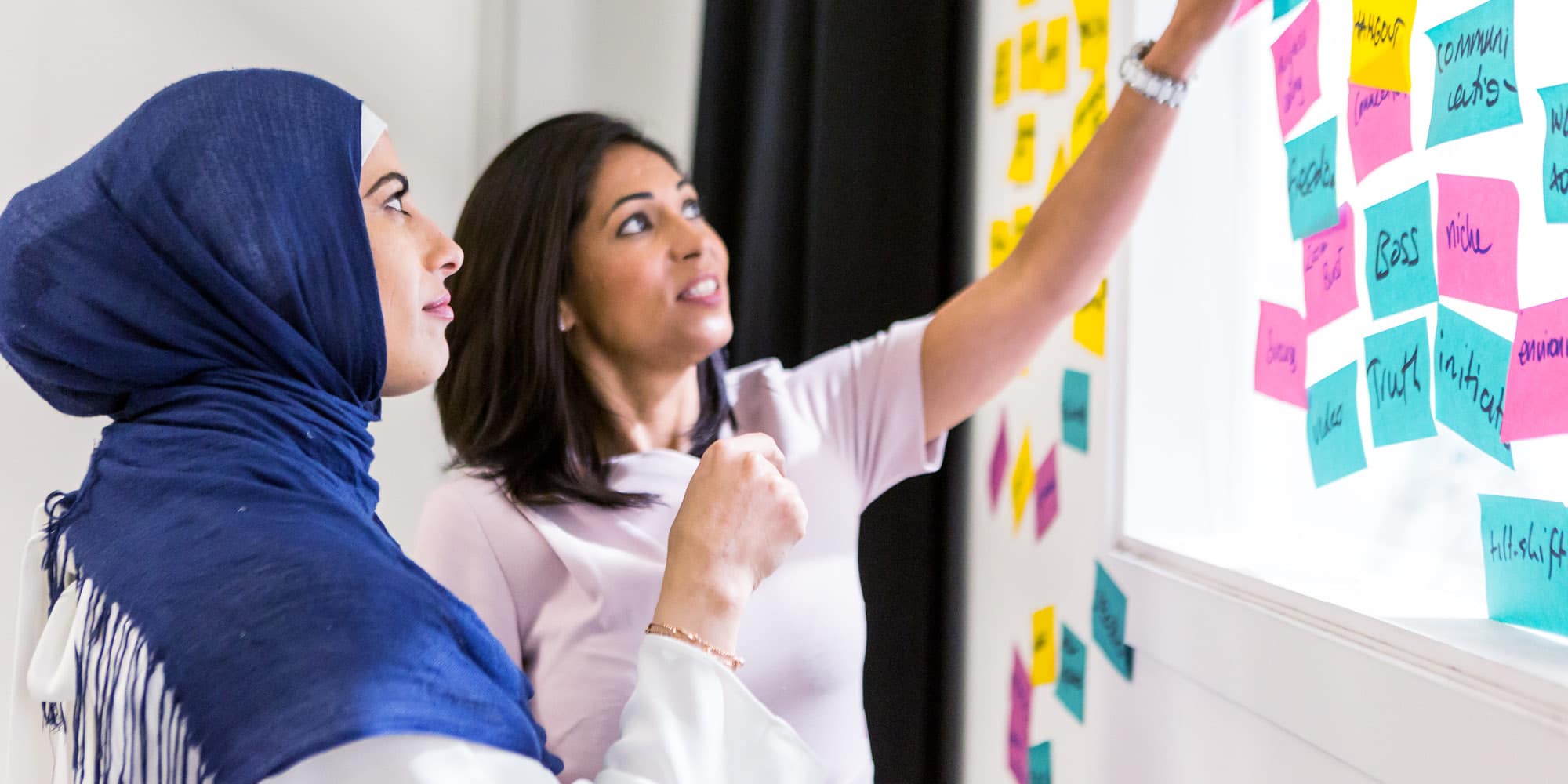Two women stand by a window covered with colorful sticky notes, discussing and pointing at the notes. In this skills training Canada session, one woman wears a blue hijab, and both appear focused and engaged in collaboration.