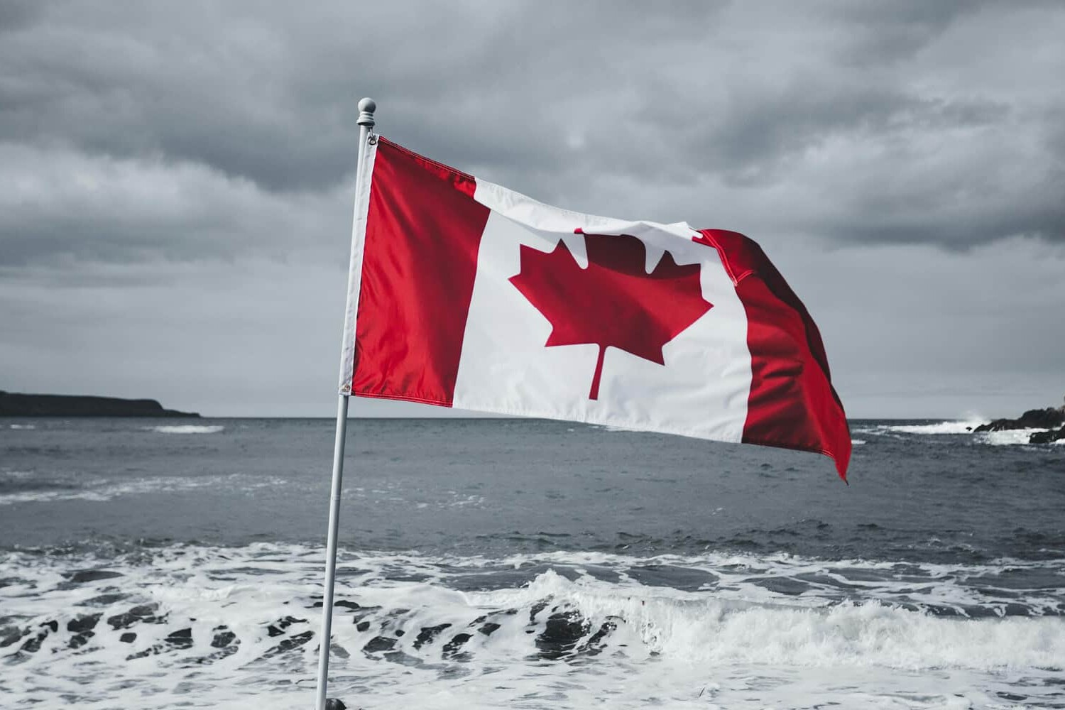 A Canadian flag with a red maple leaf waves on a pole by the ocean, with waves crashing on the shore and a cloudy, overcast sky in the background—prompting some to wonder: is one region favoured by Ottawa?.